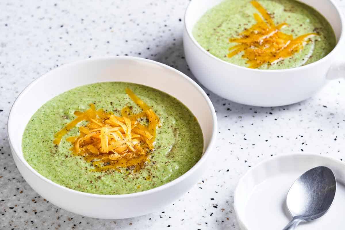 Side view of two bowls of cream of broccoli soup with grated cheese on top.
