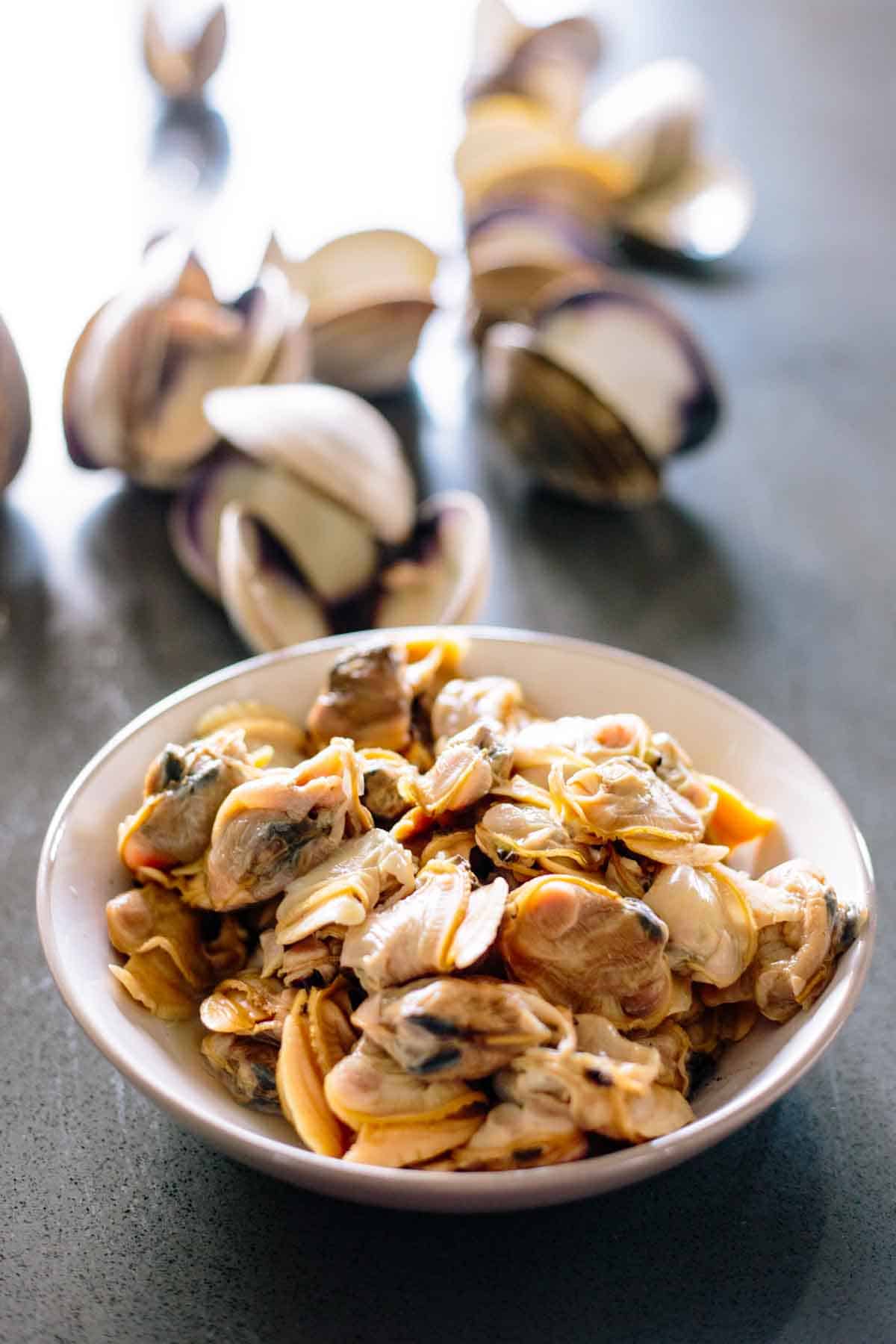 A bowl of shucked cooked clam meat with clam shells in the background.
