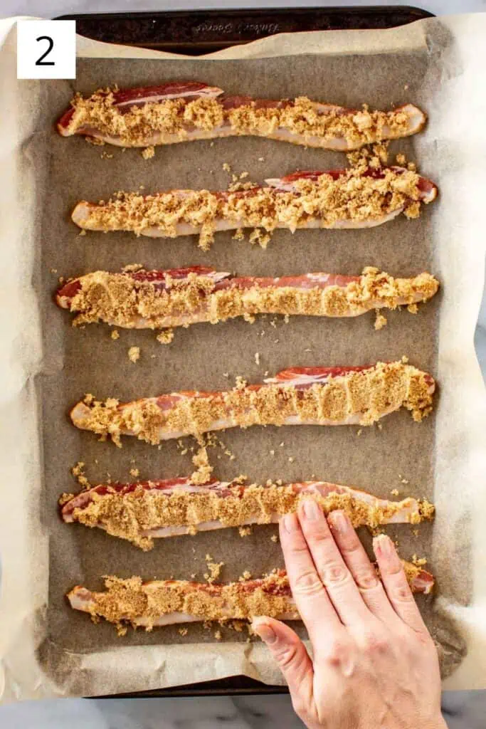 A hand pressing brown sugar into strips of bacon on a sheet pan.