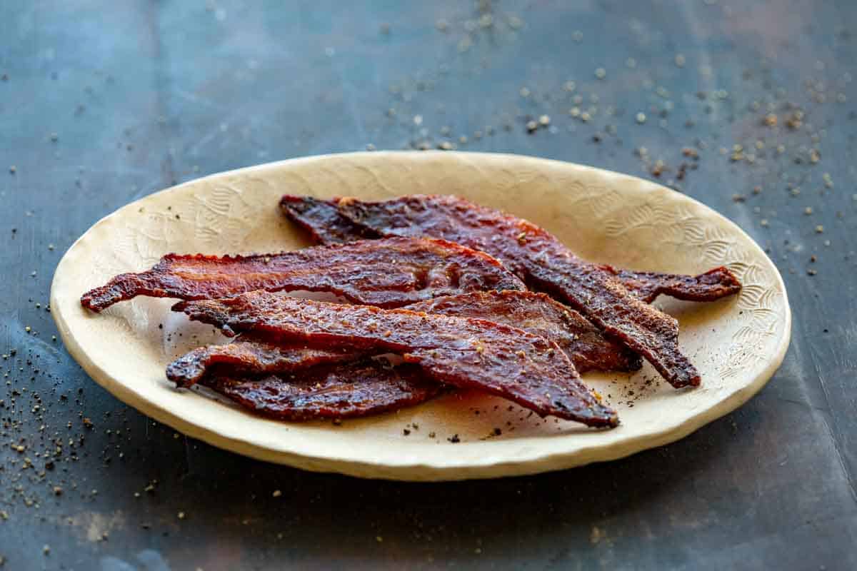 Side view of a beige oval plate with strips of brown sugar candied bacon.