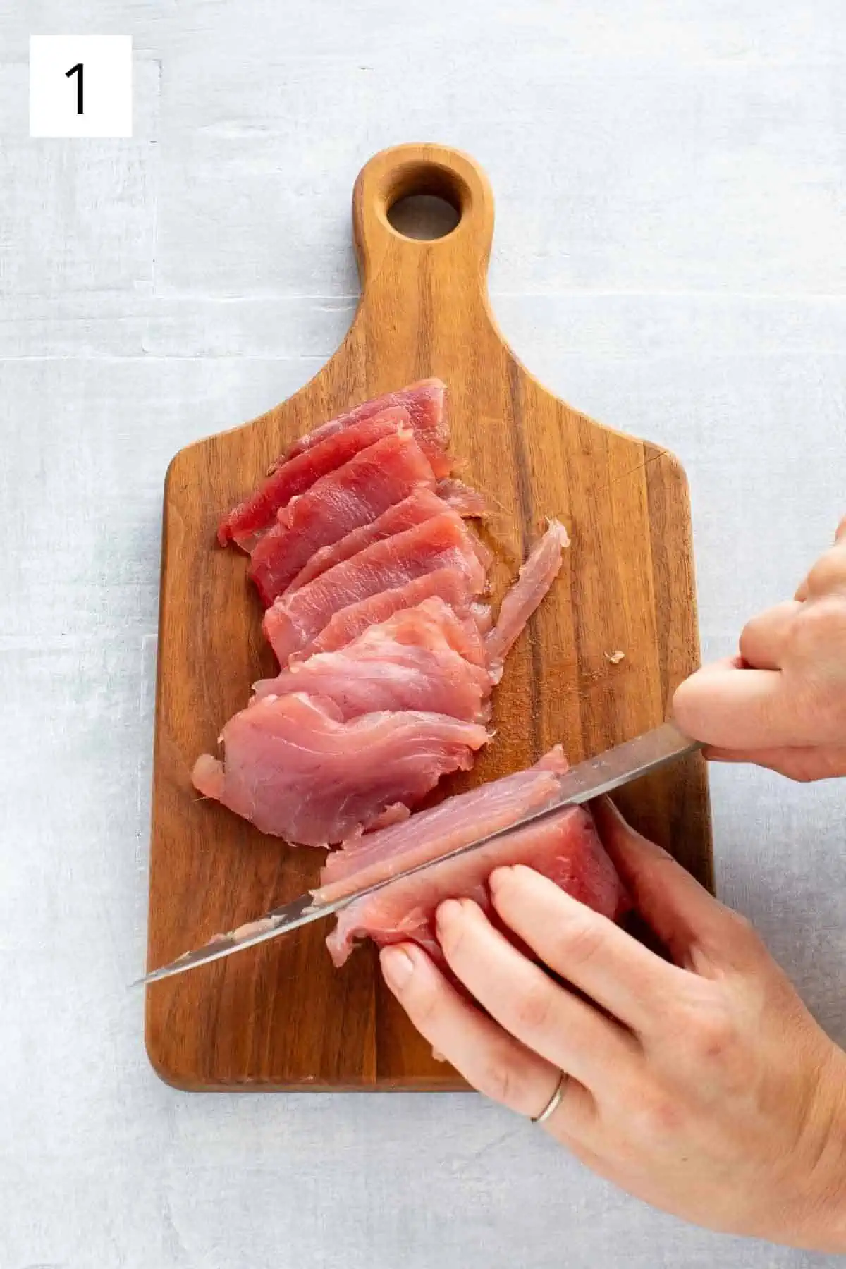 A hand thinly slicing raw tuna on a wood cutting board.