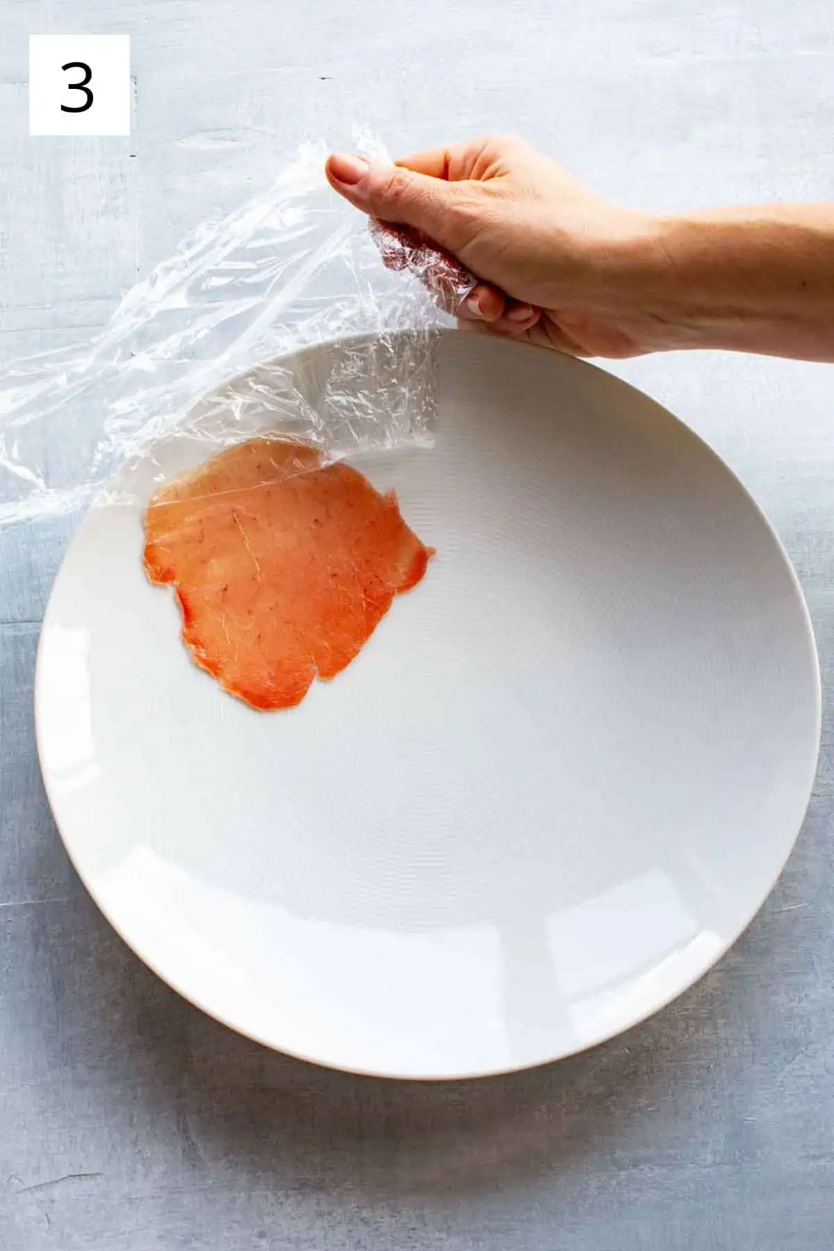 A hand removing plastic wrap from a thin slice of raw tuna on a white plate.