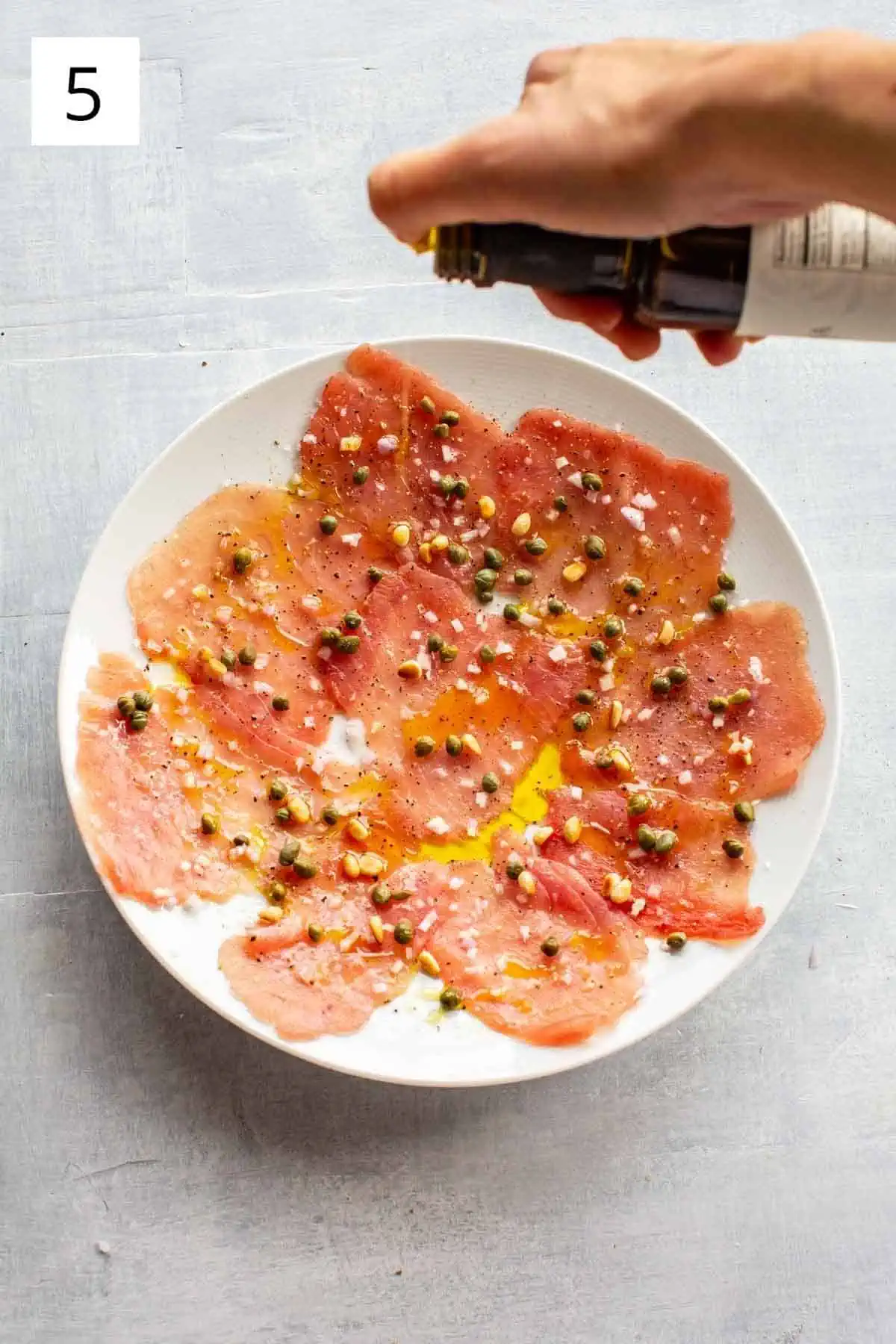 Olive oil being poured over a plate of raw tuna with capers, pine nuts and shallots.