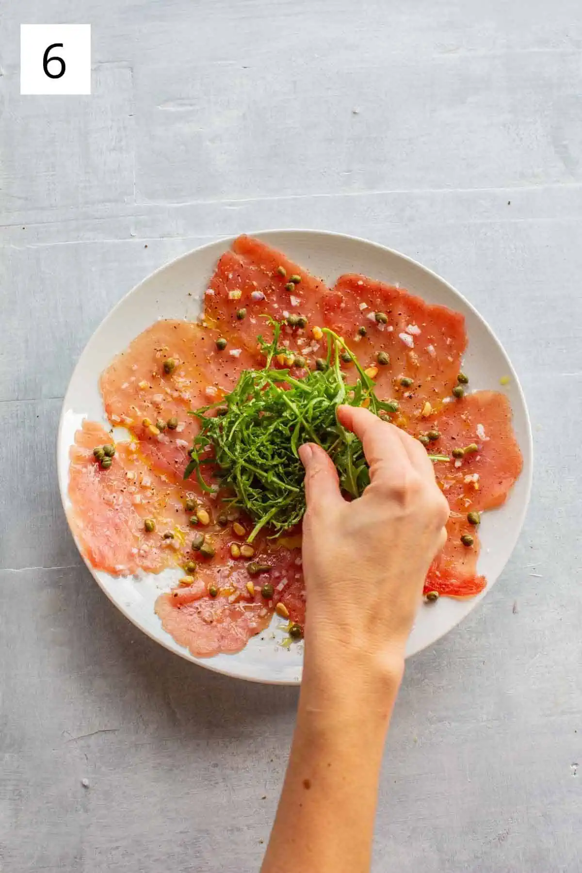A hand placing a mound of arugula over top of a plate of raw tuna.