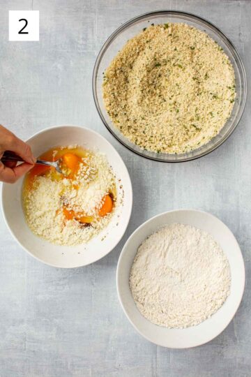 Overhead shot of a bowl of breadcrumbs, a bowl of flour and a bowl of eggs being whisked.