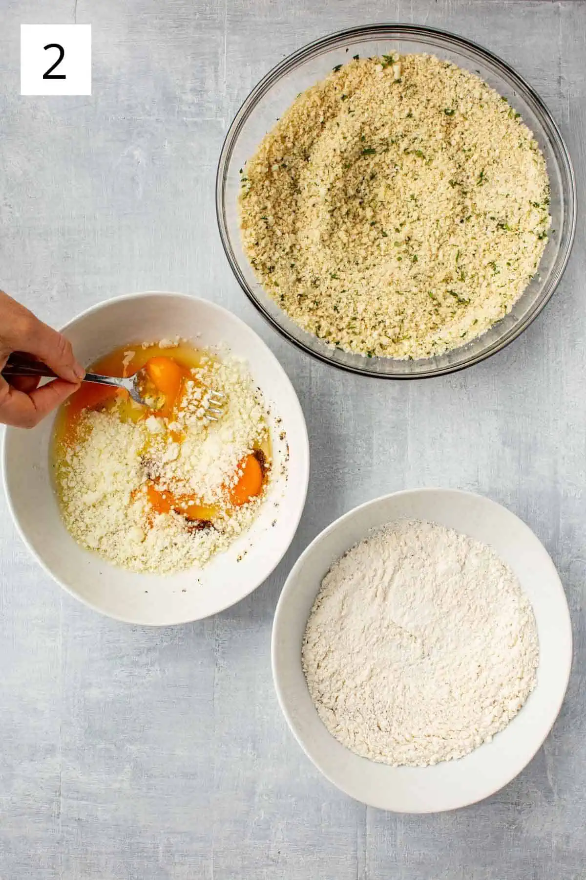 Overhead shot of a bowl of breadcrumbs, a bowl of flour and a bowl of eggs being whisked.