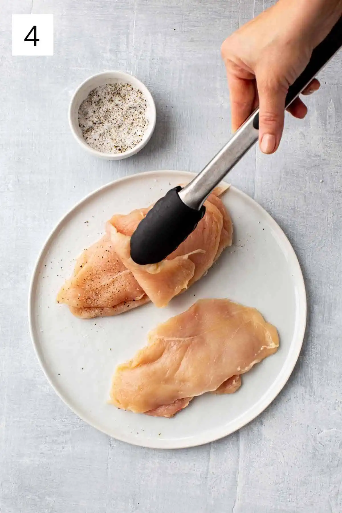 Tongs placing a raw chicken breast on to a pile of other raw chicken.