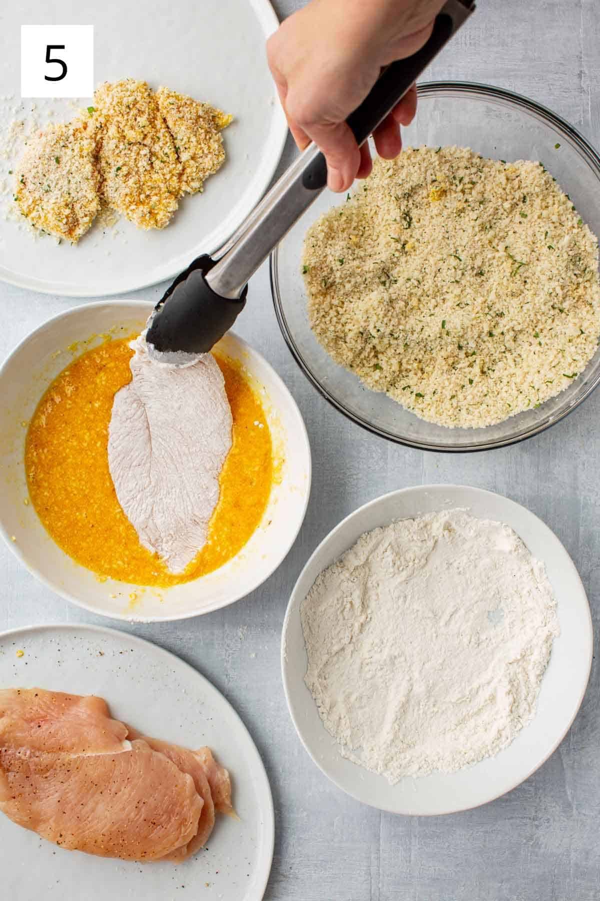 Overhead shot of a standard breading station with tongs dipping chicken into egg.