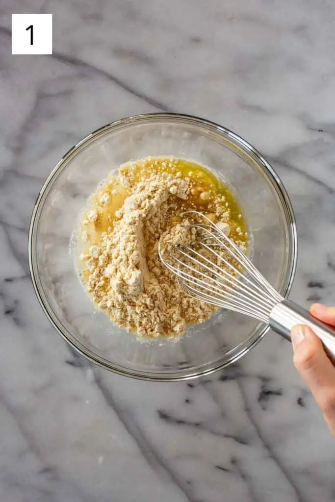 A hand whisking chickpea flour, water and olive oil together in a glass bowl.