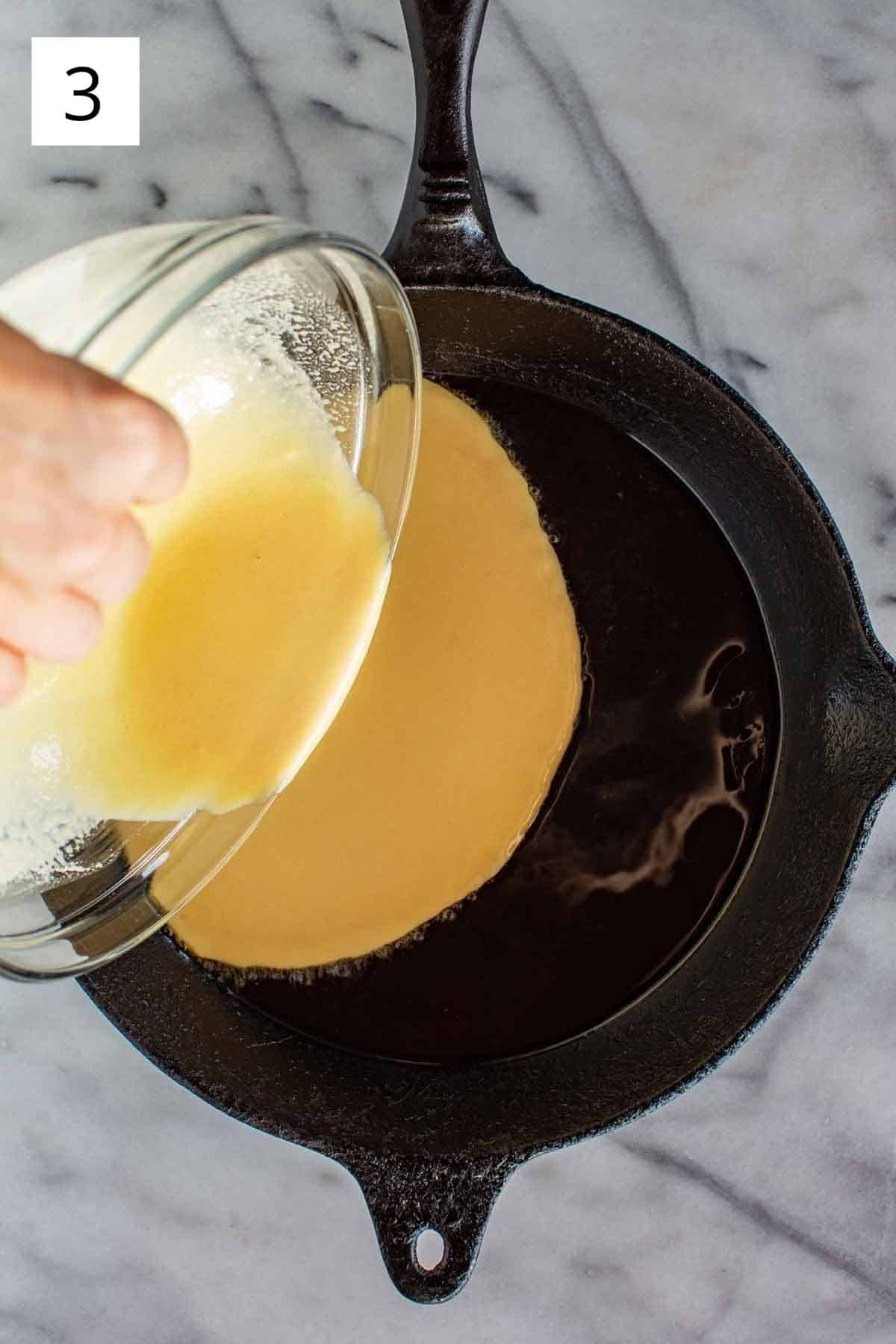 A hand pouring chickpea batter into a hot cast iron skillet.