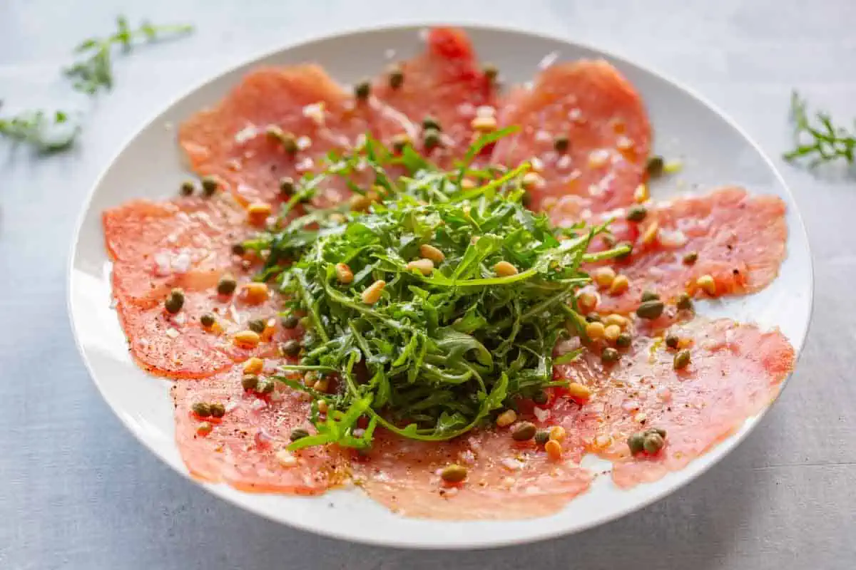 A white plate with slices of raw tuna with a mound of arugula in the center and accouterments sprinkled throughout.