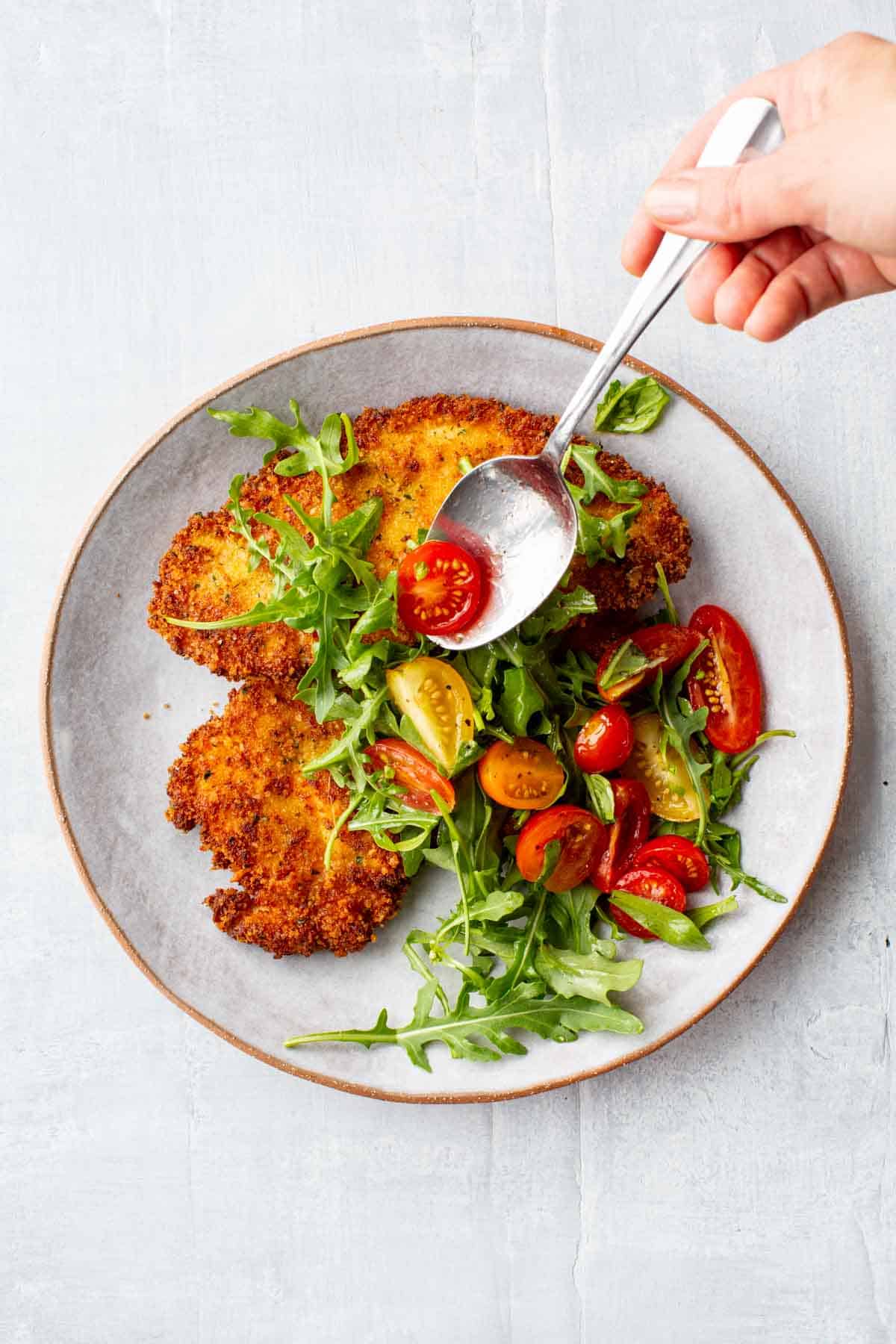 Overhead shot of tomatoes being spooned over a plate of chicken Milanese and arugula. 