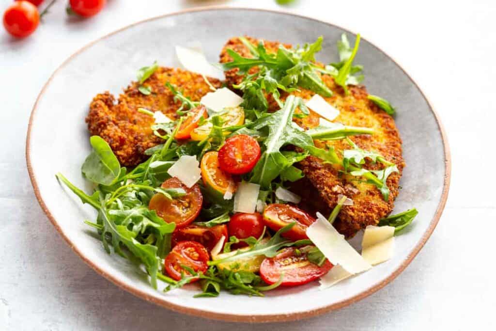 A plate of breaded fried chicken topped with fresh arugula, cherry tomato halves, and shaved parmesan cheese. The dish is placed on a white plate with a light background.