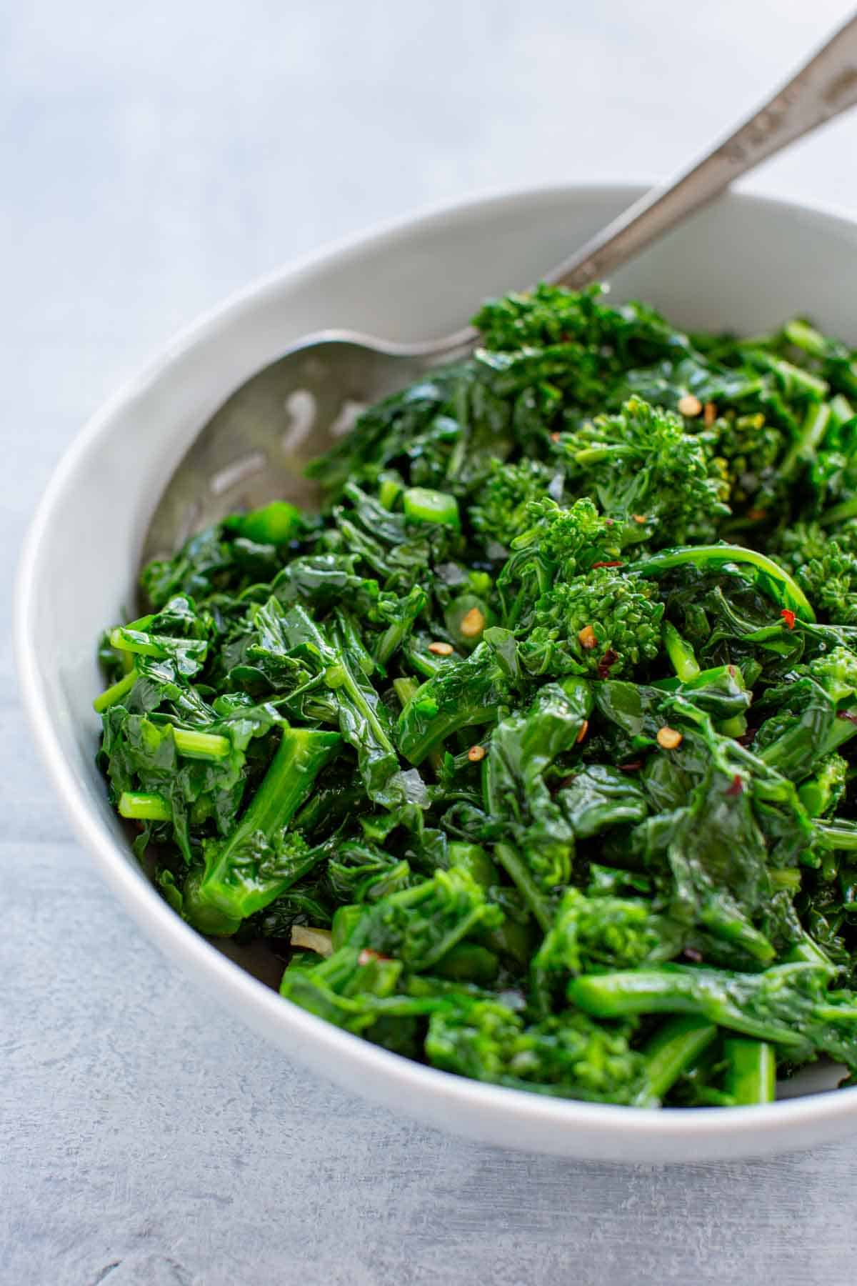 Close up of a white bowl of sautéed rapini with a serving spoon.