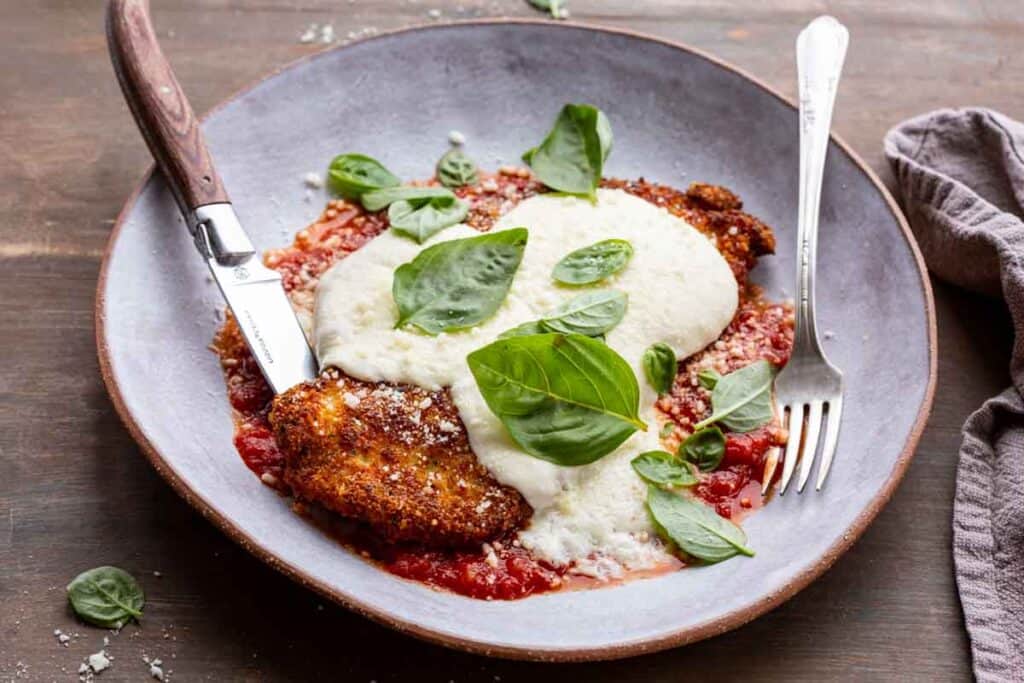 A delectable plate of chicken Parmesan is showcased, featuring a breaded chicken cutlet topped with melted mozzarella cheese, marinara sauce, and fresh basil leaves. A knife and fork are placed beside the dish, ready for use.