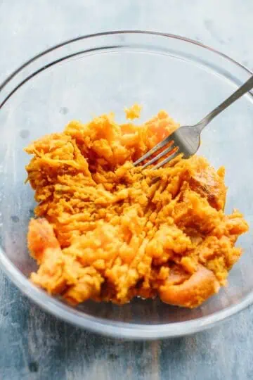 Skinless baked sweet potatoes being mashed with a fork in a glass bowl.
