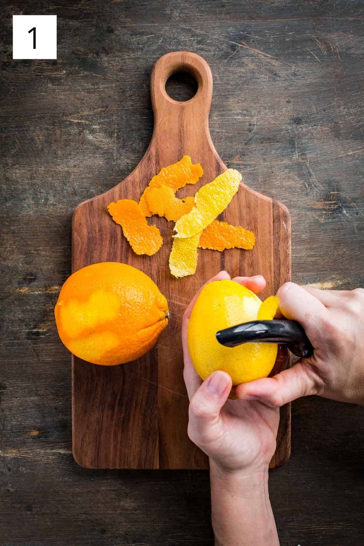 A hand removing citrus zest from a lemon with a peeler.