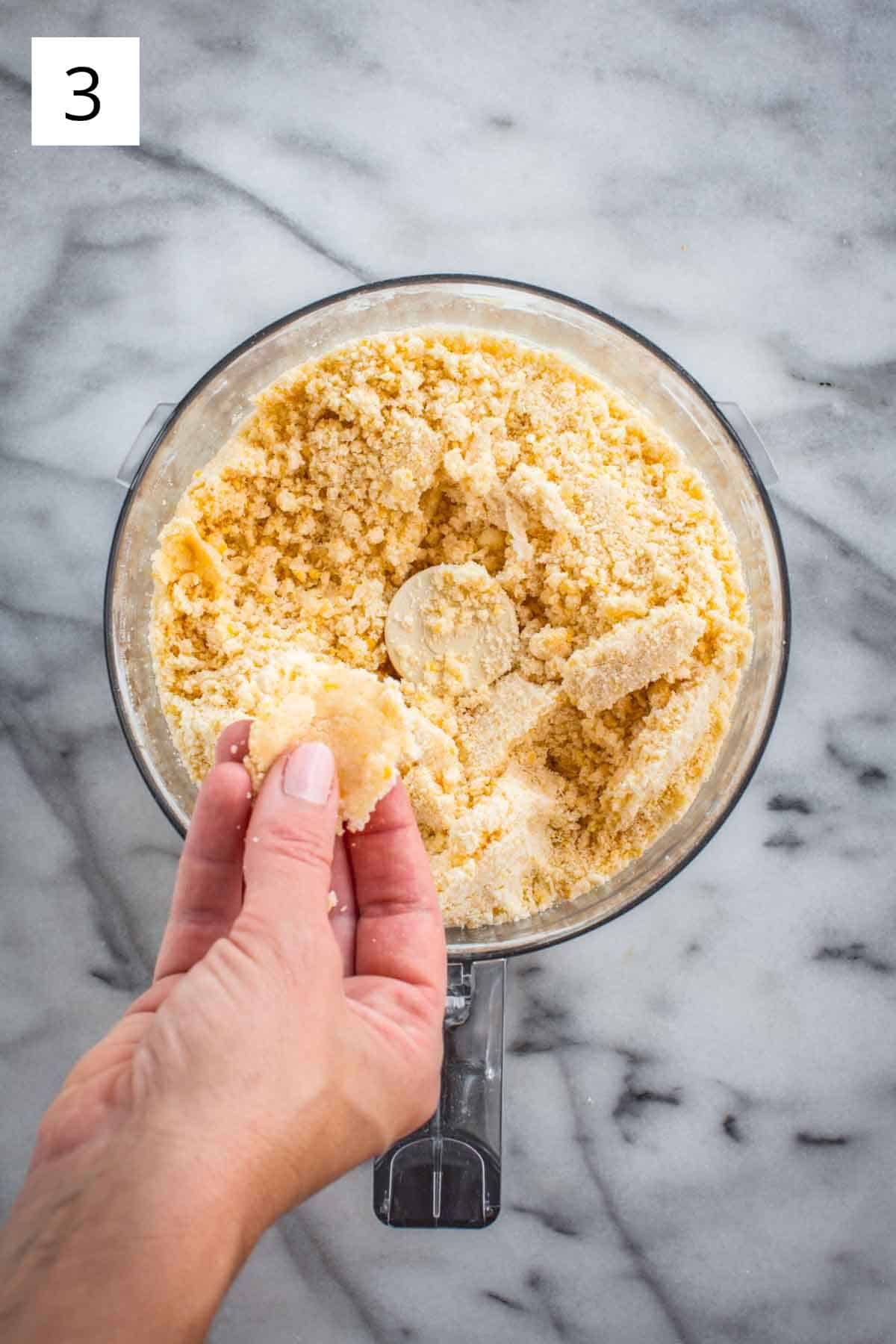 A hand squeezing a bit of dough together over a food processor.