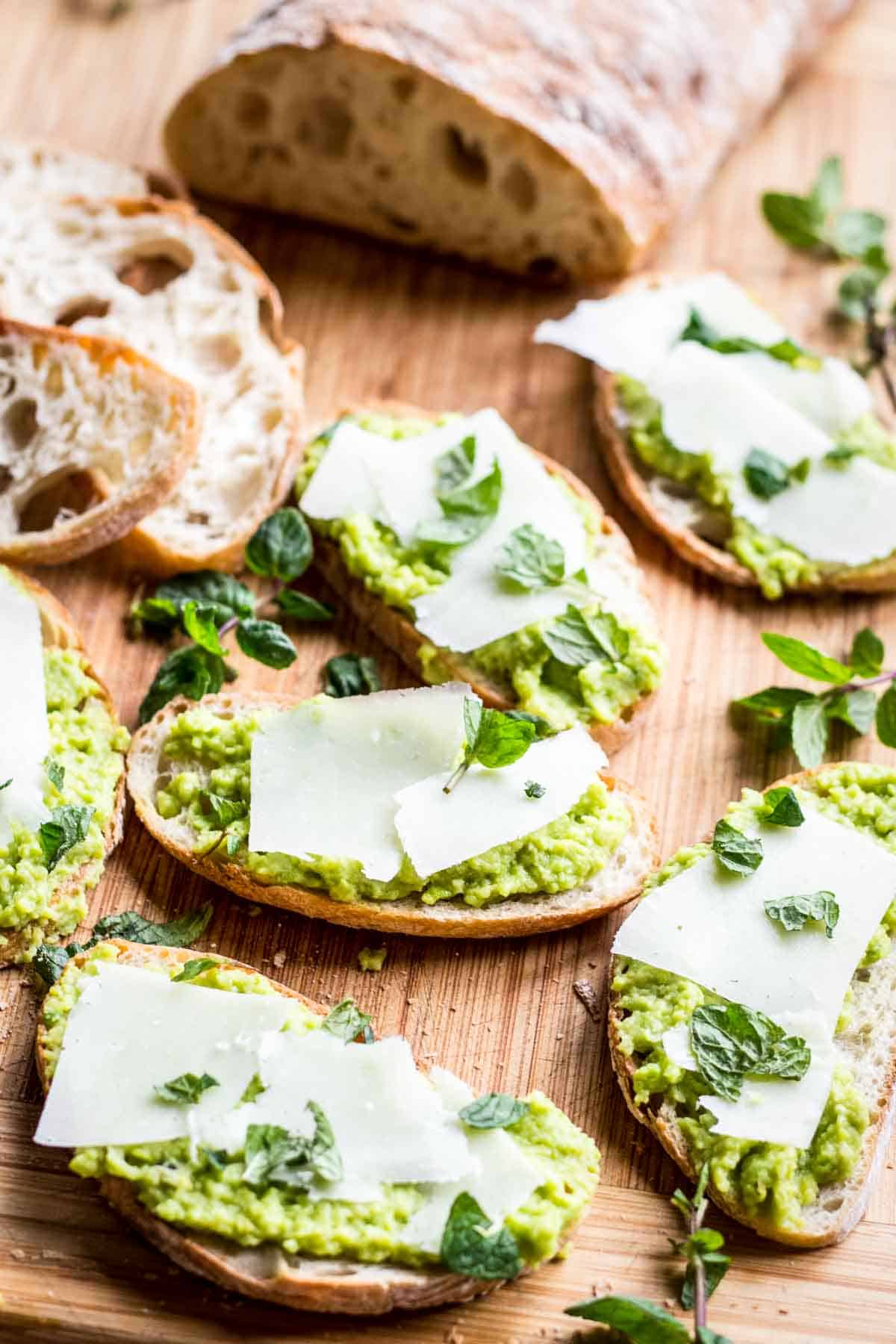 A wooden board with pieces of toast, Fava bean spread, slices of cheese and mint top.