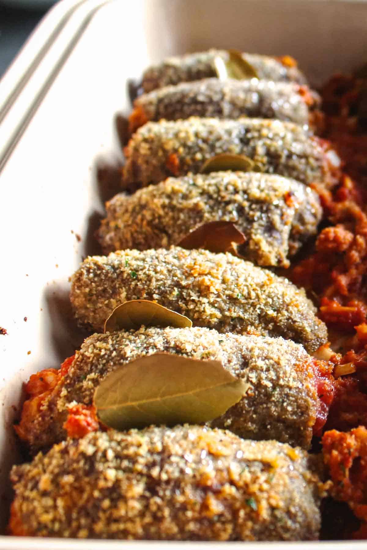 Breaded beef roll ups baked in a pan with bay leaves tucked in between each.