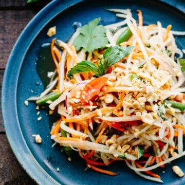 Thai papaya salad with different colored vegetables and herbs on a blue plate.