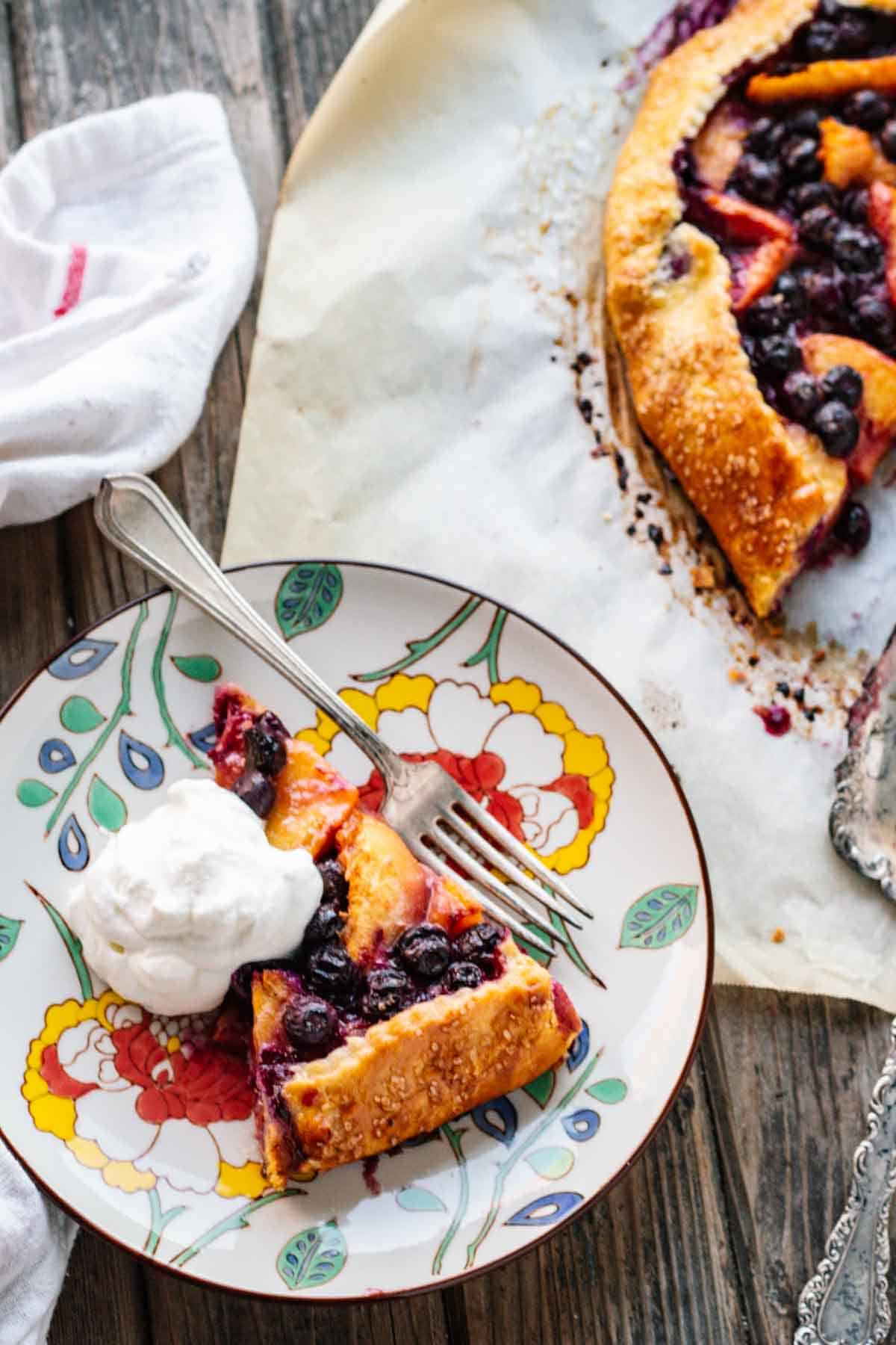 A slice of blueberry peach crostata on a plate with whipped cream and a fork.
