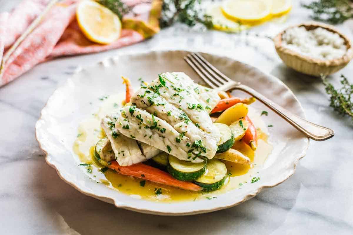 Butter poached fish over vegetables on a white plate with a fork.