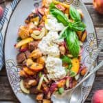 A serving dish of grilled peach and bread salad with burrata and serving spoons.
