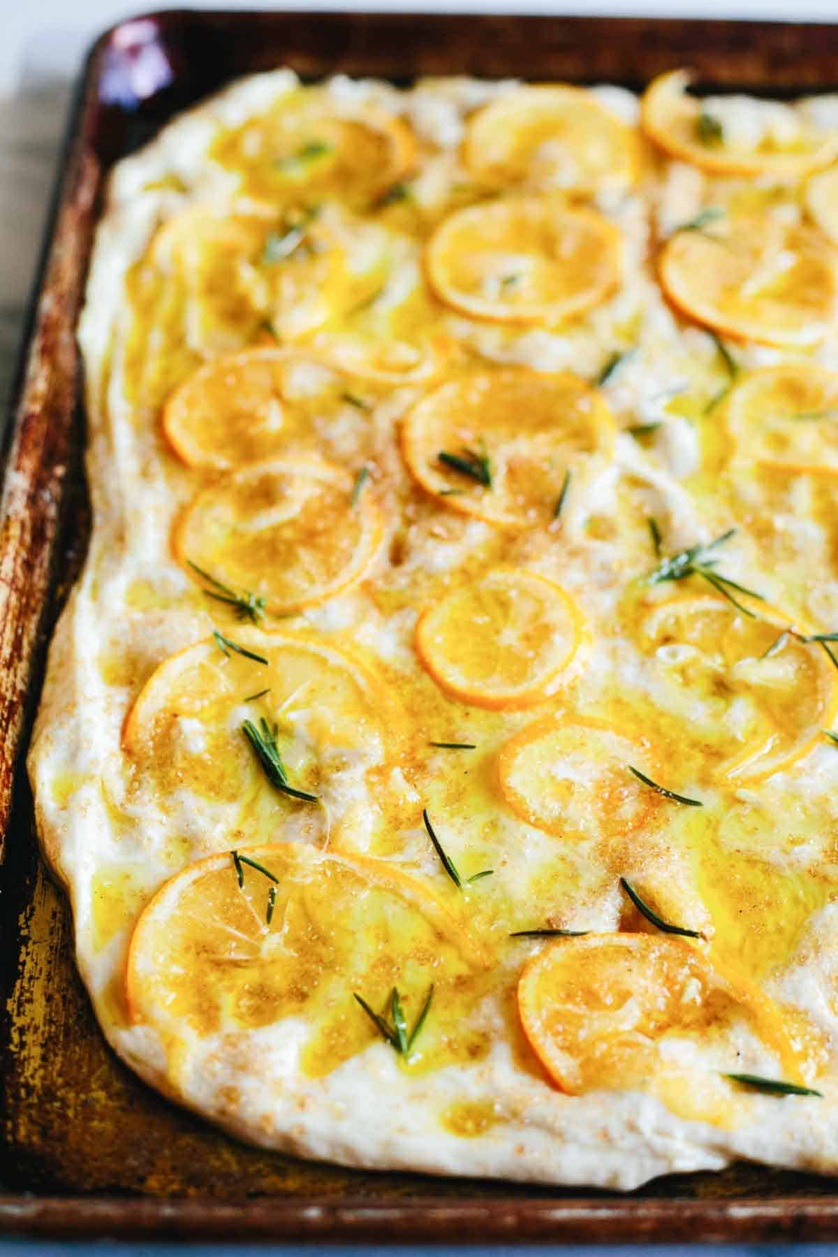 Focaccia dough spread out on a baking pan with sliced lemons, rosemary and raw sugar on top.