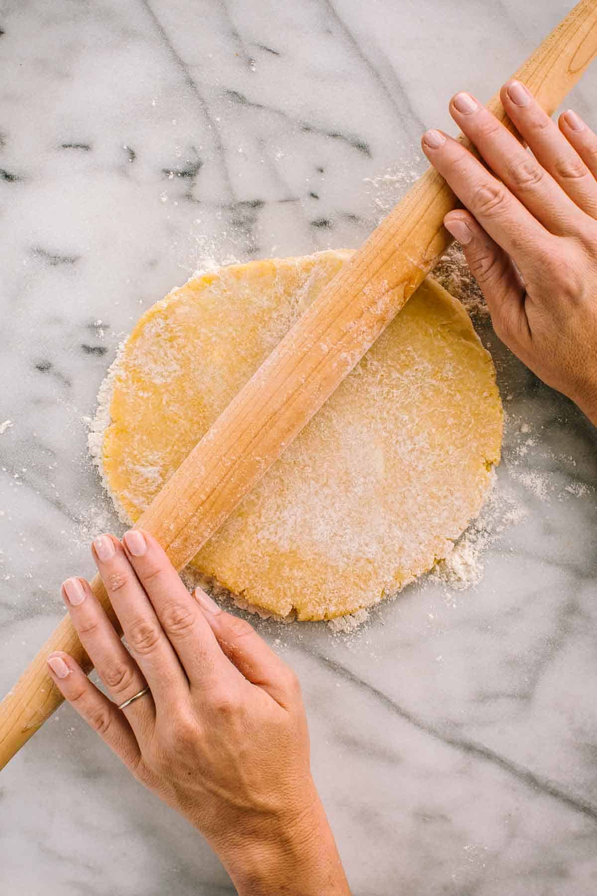 Hands rolling out a piece of pie dough with a French style tapered rolling pin. 