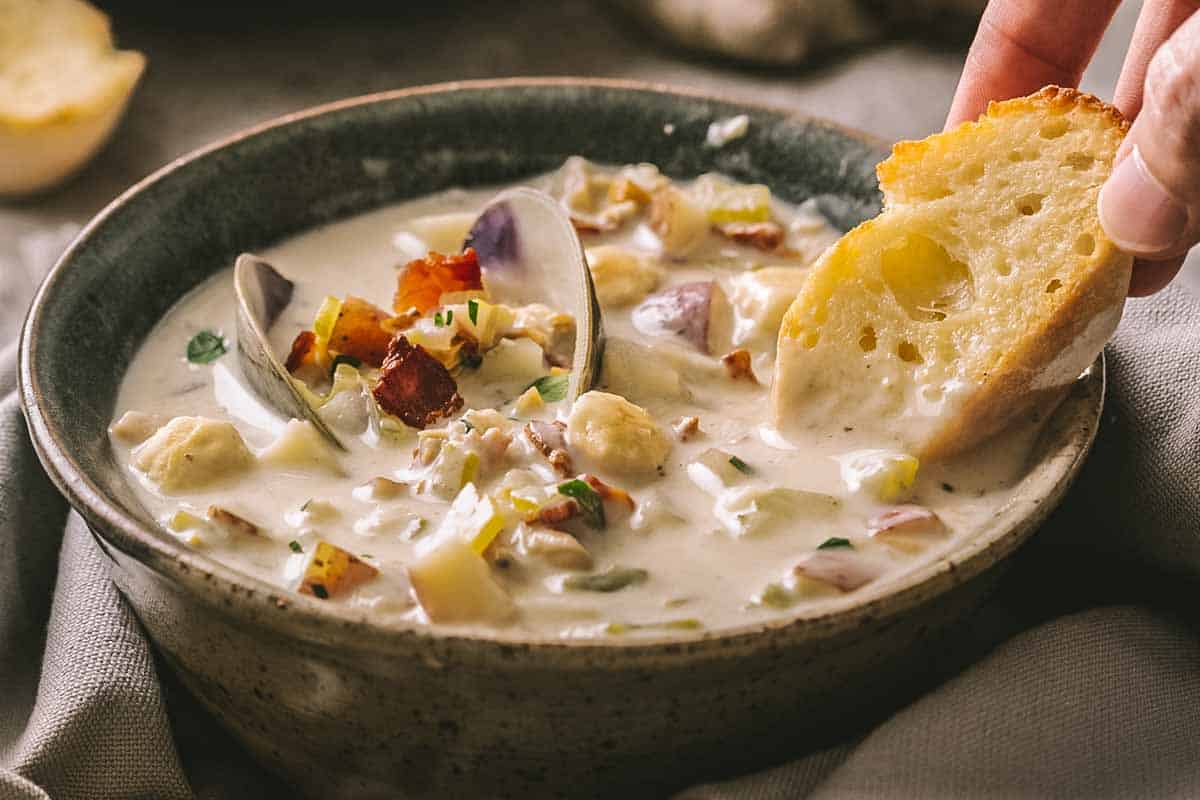 A hand dipping bread into a bowl of clam chowder