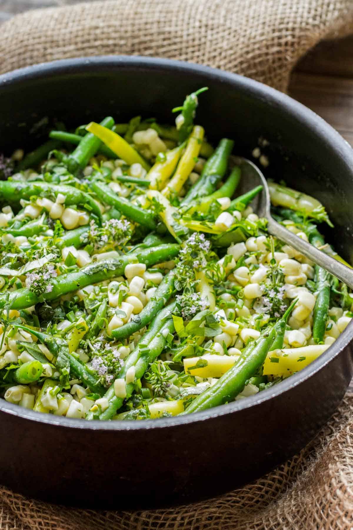 A spoon in a pan of green beans with white corn and herb butter.