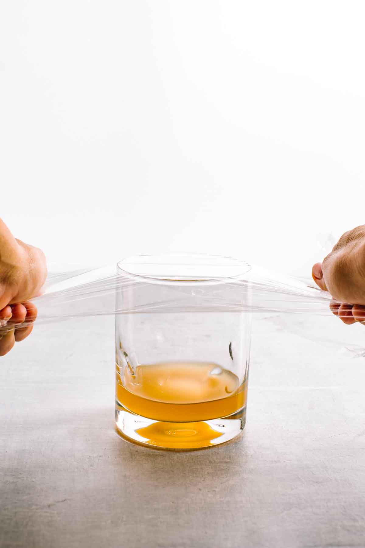 Hands placing plastic wrap on a glass filled with amber liquid.