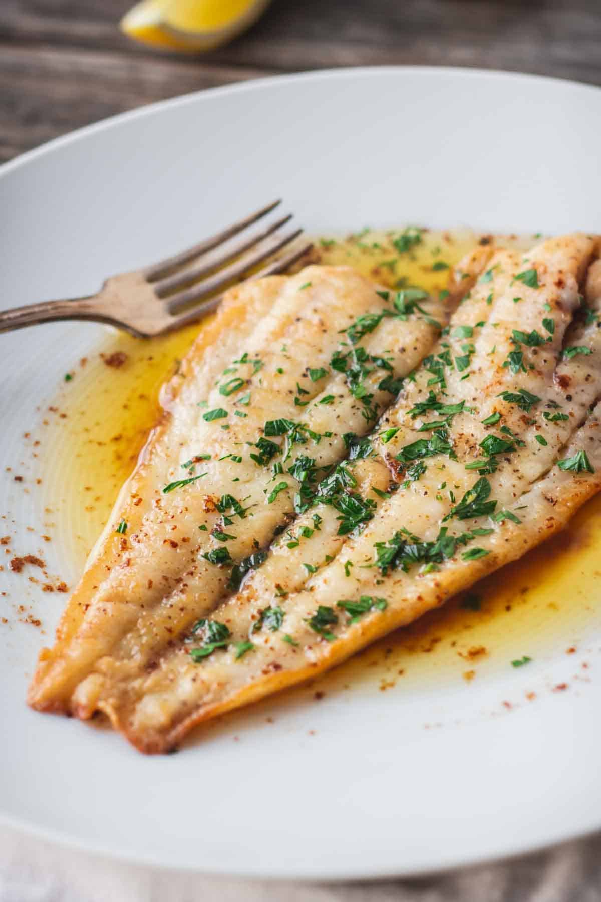 A fillet of flounder on a white plate with browned butter, parsley and a fork.