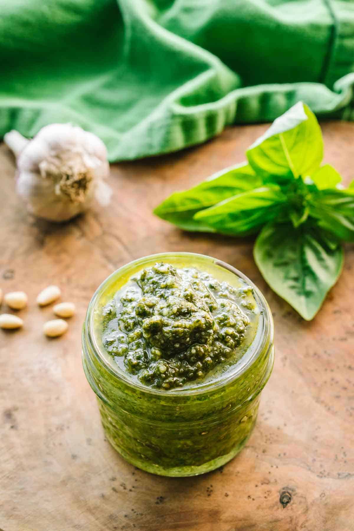 A small jar of pesto with a bulb of garlic, pine nuts and a sprig of basil. 