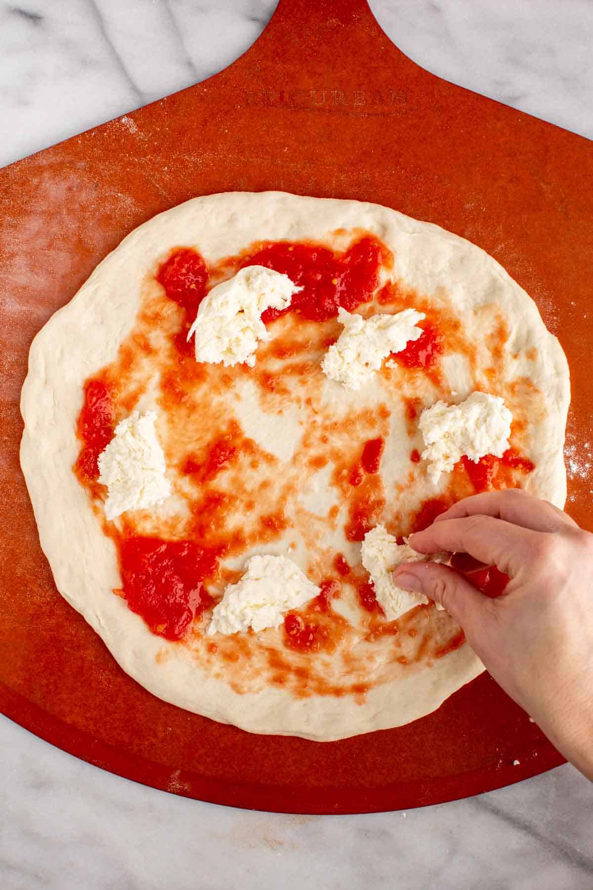 A hand placing torn pieces of fresh mozzarella cheese on pizza dough.