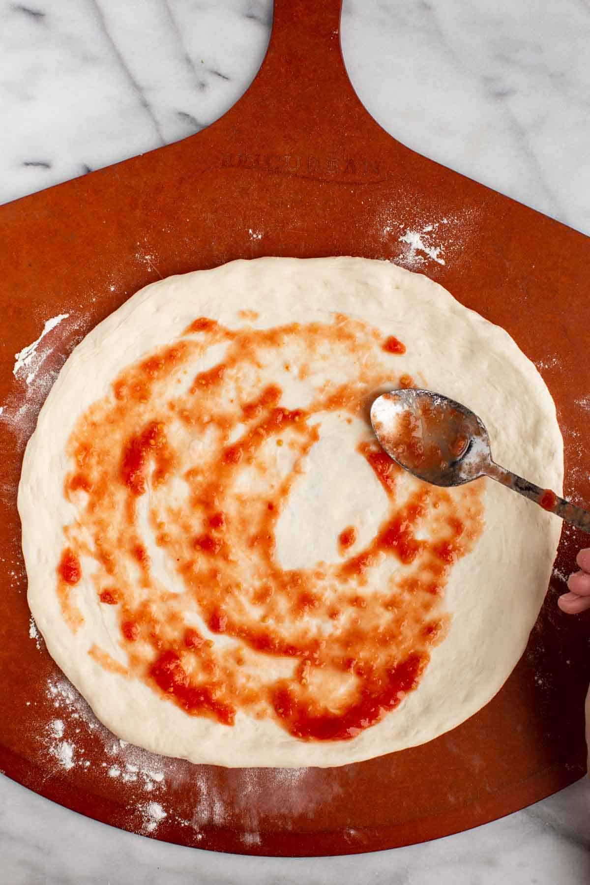 A spoon spreading pizza sauce on stretched out dough.