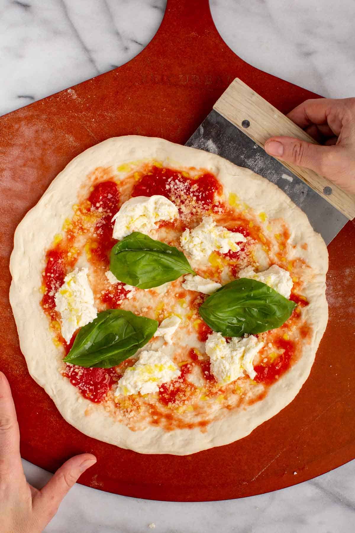 A hand using a bench scraper to loosen an uncooked pizza on a peel.