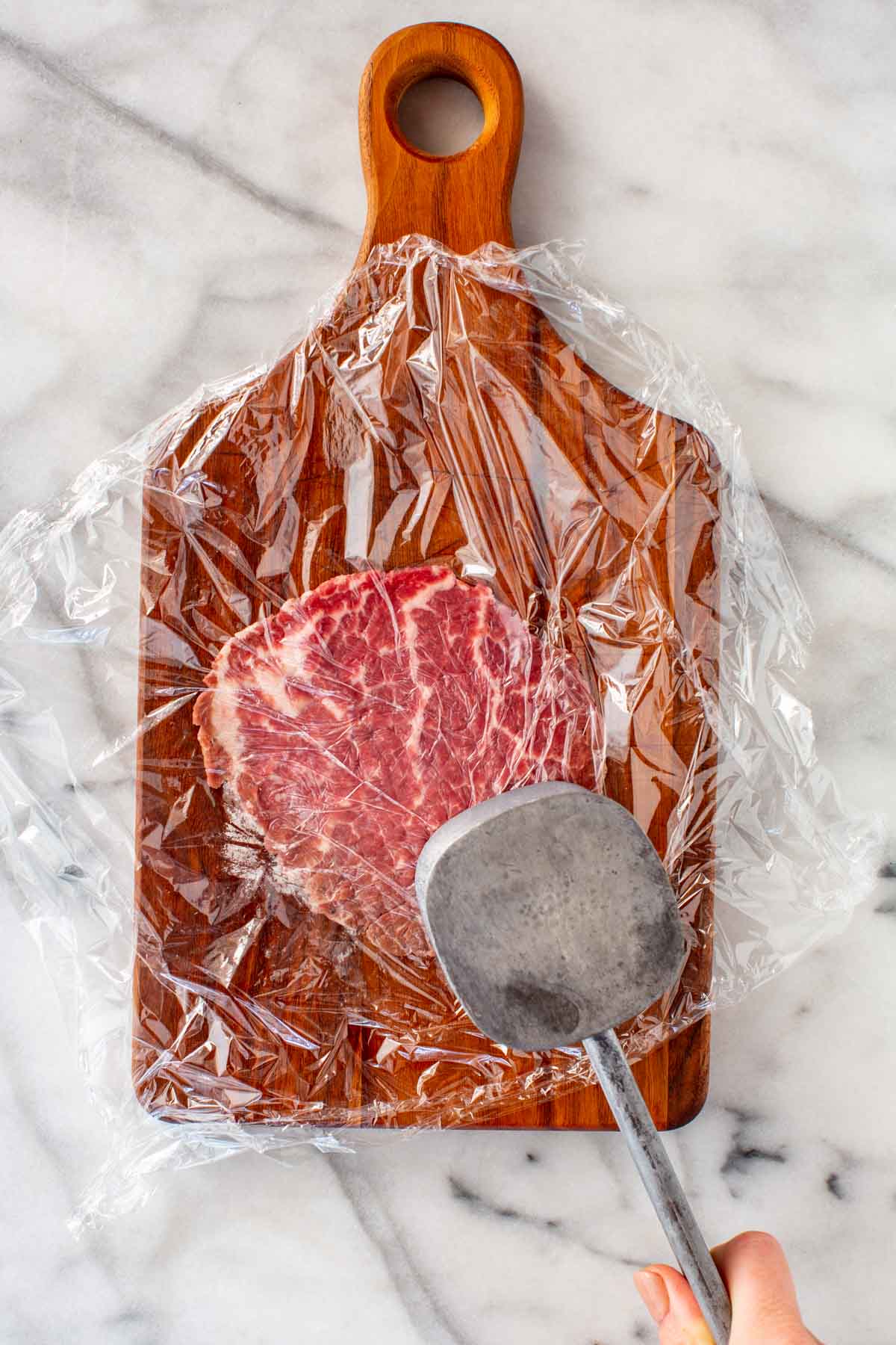 Pounding out a slice of beef with a meat mallet under a sheet of plastic wrap.