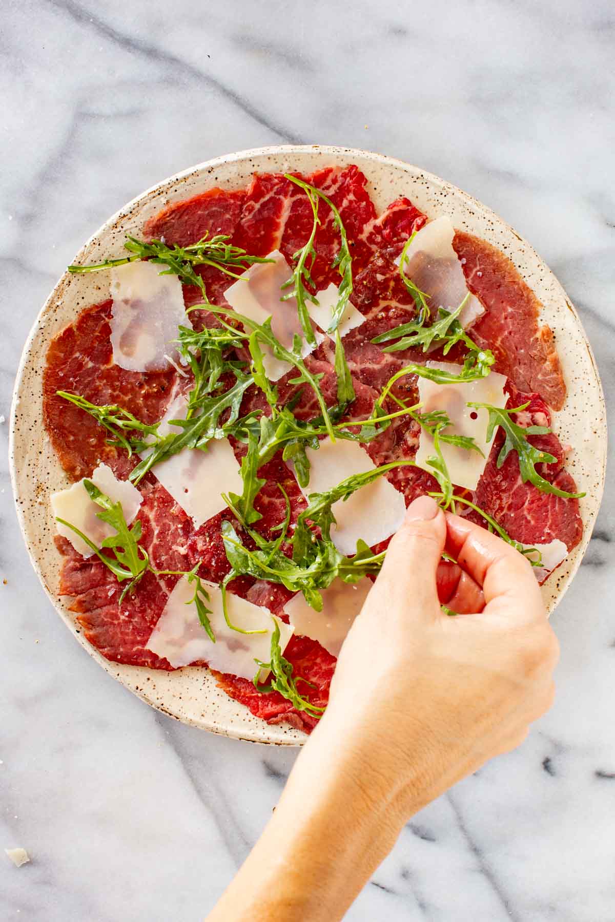 Adding shaved parmesan and arugula to a plate of beef carpaccio.