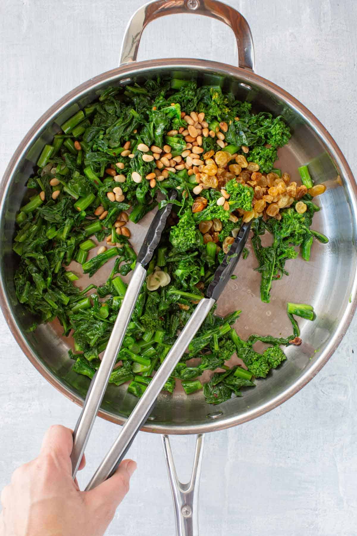 Mixing pine nuts and raisins into sautéed broccoli rabe in a pan.