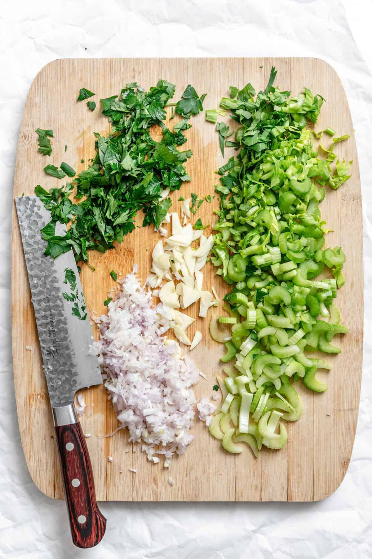 Chopped celery, shallots and parsley on a cutting board with a chefs knife.