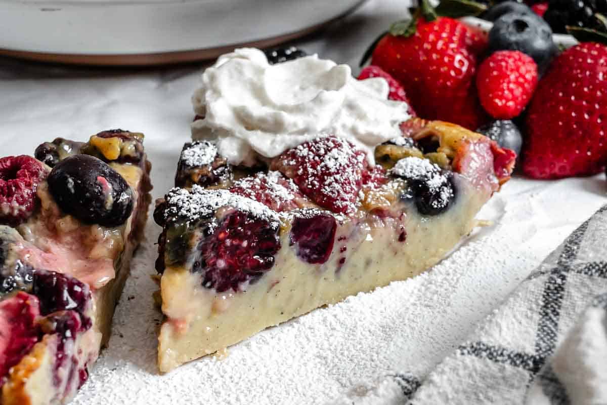 Slice of berry clafoutis on a plate with whipped cream and fresh berries, dusted with powdered sugar.