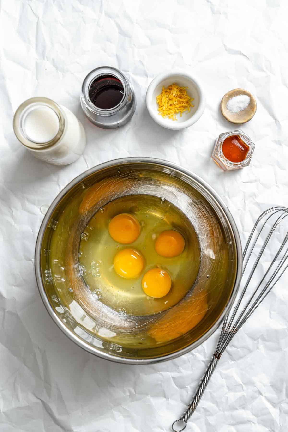 Four eggs in a metal mixing bowl with ingredients in small containers nearby including milk, vanilla, lemon zest, salt, and maple syrup.