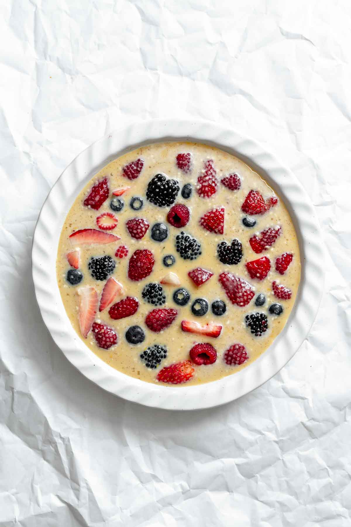 Mixed berries added to a custard mixture in a white ceramic pie dish.