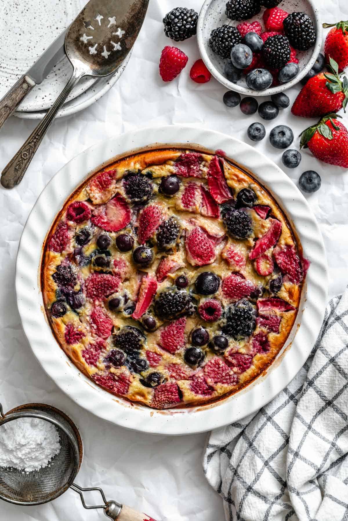 Baked berry clafoutis in a white ceramic dish with golden edges and colorful berries throughout.
