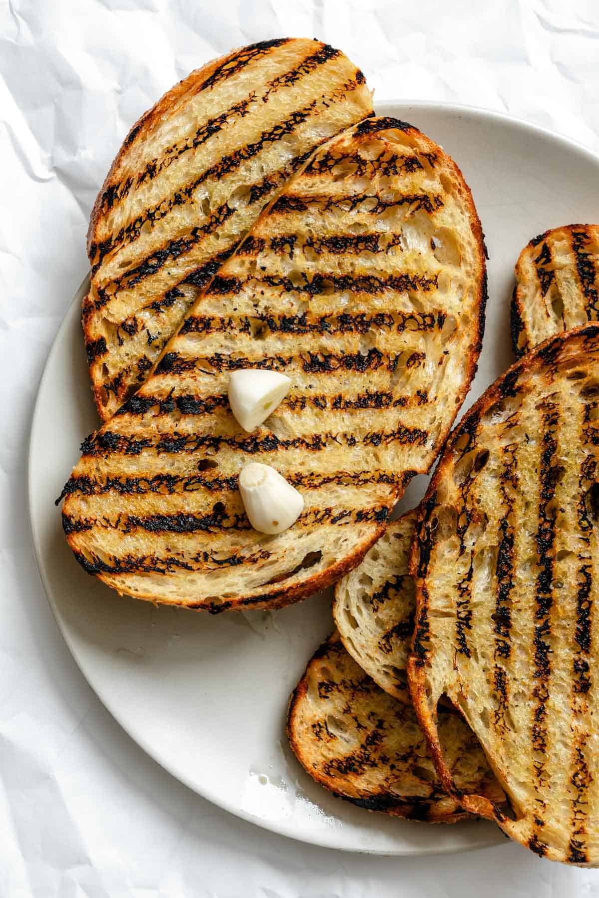 A white plate with grilled bread slices showing distinctive grill marks and two garlic cloves placed on top of one slice.