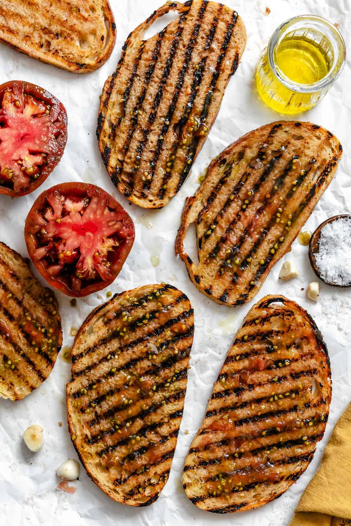 Overhead view of several pan con tomate slices with sliced tomatoes, a small jar of olive oil, and a dish of salt.