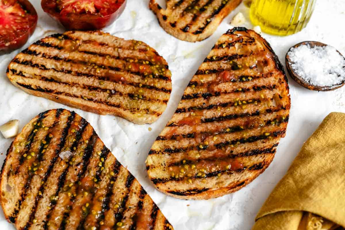 More grilled bread slices on parchment paper with tomatoes, olive oil, and a jar of olive oil in the corner.
