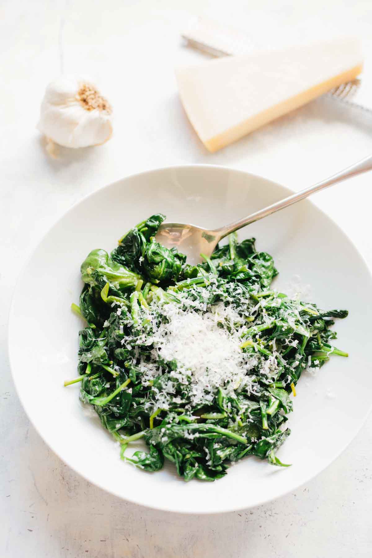 A white bowl of sautéed spinach topped with freshly grated Parmigiano-Reggiano cheese and a silver spoon, with a head of garlic and slice of cheese visible in the background.